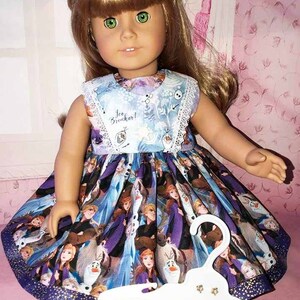 K&ouml;nnte beinhalten: Eine Puppe tr&auml;gt ein &auml;rmelloses Kleid mit blauem Oberteil und einem Rock mit Figuren aus dem Film "Die Eisk&ouml;nigin". Das Kleid hat eine wei&szlig;e Spitzentrimmung und die Worte "Ice Breaker!" sind auf dem Oberteil aufgedruckt. Ein wei&szlig;er Kleiderb&uuml;gel befindet sich vor der Puppe.