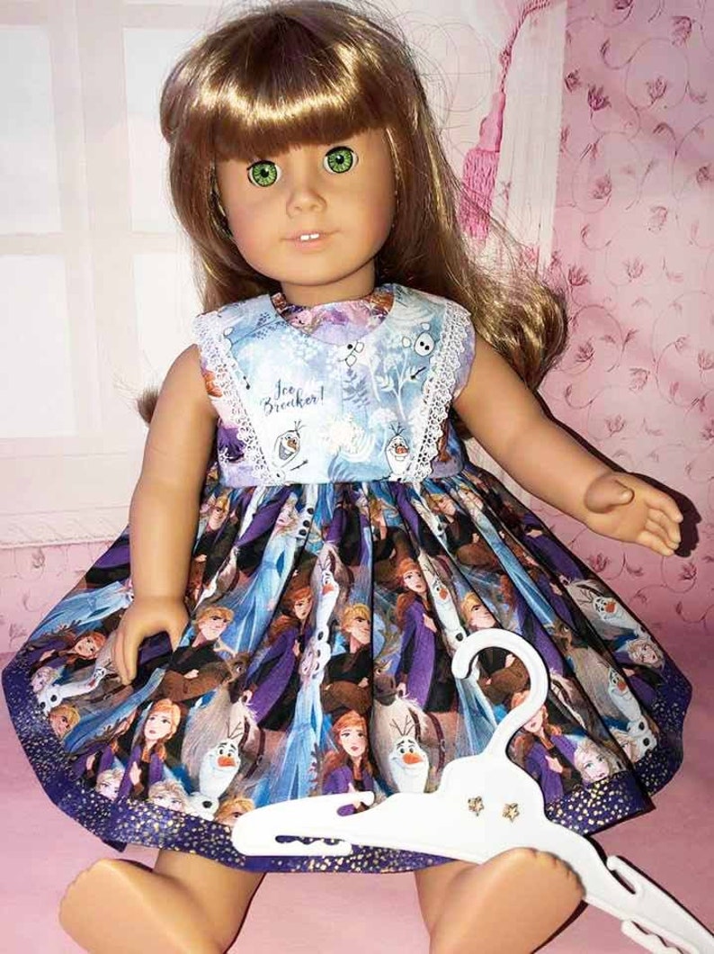 K&ouml;nnte beinhalten: Eine Puppe tr&auml;gt ein &auml;rmelloses Kleid mit blauem Oberteil und einem Rock mit Figuren aus dem Film "Die Eisk&ouml;nigin". Das Kleid hat eine wei&szlig;e Spitzentrimmung und die Worte "Ice Breaker!" sind auf dem Oberteil aufgedruckt. Ein wei&szlig;er Kleiderb&uuml;gel befindet sich vor der Puppe.