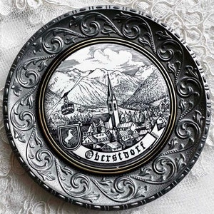 May include: Decorative metal plate with a detailed black and white illustration of a mountain village, a church, and a cable car. The plate has a silver-colored, ornate border and the word "Oberstdorf" is printed on the plate.