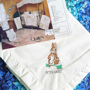 May include: A white receiving blanket with a Peter Rabbit embroidery. The blanket has a satin binding and is made of soft, plush fabric. The image shows a crib with a canopy and other baby bedding items.