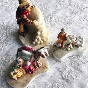 May include: Three ceramic figurines depicting a nativity scene. The first figurine shows a man in a brown and red outfit rolling a snowball. The second figurine shows a man and a woman in brown and green outfits with a flock of white sheep. The third figurine shows a man in a blue and red outfit riding a camel with a woman in a red and green outfit.