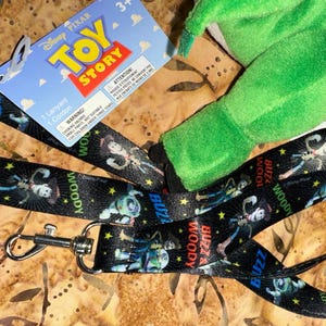 May include: Black lanyard featuring characters Buzz Lightyear and Woody from Disney Pixar's Toy Story. The lanyard has a metal clasp and a blue and red Toy Story tag. A green plush toy is also visible.