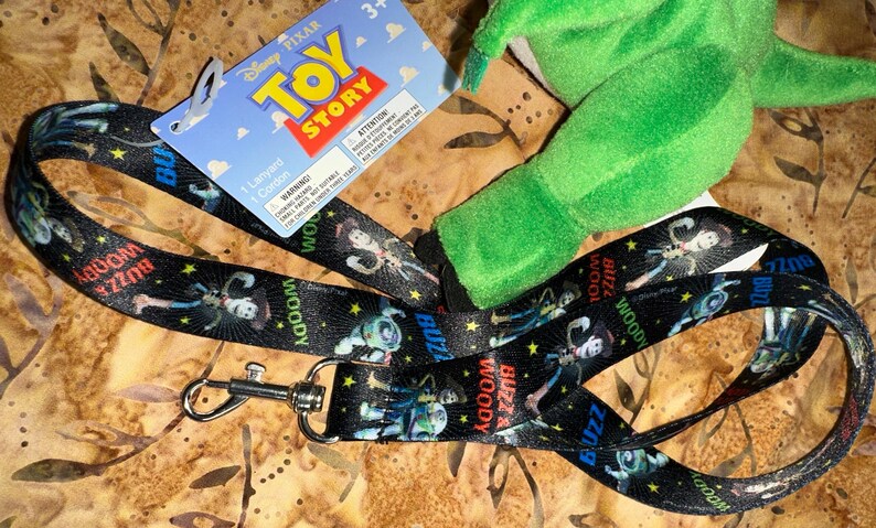 May include: Black lanyard featuring characters Buzz Lightyear and Woody from Disney Pixar's Toy Story. The lanyard has a metal clasp and a blue and red Toy Story tag. A green plush toy is also visible.