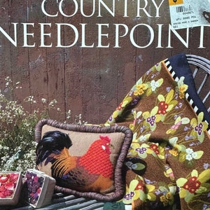 May include: A needlepoint pillow featuring a rooster with red, orange, and brown feathers. The pillow is on a wooden bench with a brown and green needlepoint throw with floral designs. The text "AMERICAN COUNTRY NEEDLEPOINT" and "JIM WILLIAMS" are visible on the image.