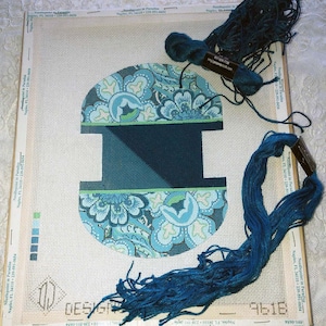 May include: Needlepoint canvas with a floral design in shades of blue and teal, featuring the letter "I". Several skeins of blue embroidery floss are arranged around the canvas, ready for stitching. The canvas is on a wooden frame.