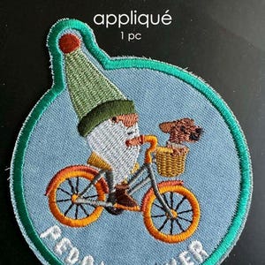 May include: Embroidered appliqué patch featuring a gnome riding an orange bicycle with a dog in the basket. The patch has a light blue background with the words "PEDAL POWER". The patch is outlined in green.