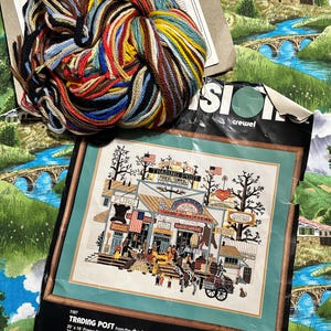 May include: A cross-stitch kit featuring a detailed illustration of a trading post, with a colorful skein of yarn on top. The kit includes the text "TRADING POST" and "1167". The kit measures 20 x 16 inches without a mat.