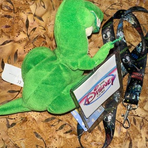 May include: A green plush toy dinosaur, Rex from Toy Story, with a Disney Store tag and a black lanyard featuring Toy Story characters. The lanyard has a silver clasp. The background has a brown and tan pattern.
