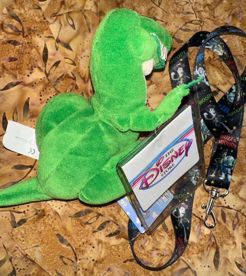 May include: A green plush toy dinosaur, Rex from Toy Story, with a Disney Store tag and a black lanyard featuring Toy Story characters. The lanyard has a silver clasp. The background has a brown and tan pattern.