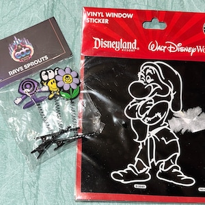 May include: Three Rave Sprouts hair clips featuring a purple swirl, a yellow smiley face, and a purple flower, each on a black metal spring. Beside them is a Disneyland and Walt Disney World vinyl window sticker depicting a white Grumpy character.