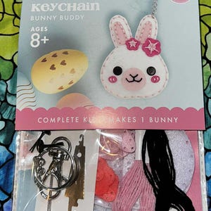 May include: A felt keychain kit featuring a white bunny with pink accents and a flower. The kit includes materials to make one bunny, with the text "FELT KEYCHAIN BUNNY BUDDY AGES 8+". The kit also includes a keychain, thread, and other accessories.