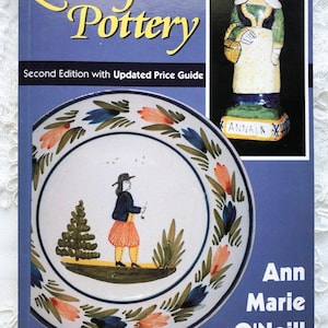 May include: Book cover for "Quimper Pottery" by Ann Marie O'Neill, second edition with updated price guide. The cover features a blue and white plate with a painted scene of a person in traditional clothing, a tree, and flowers. The book title is in yellow lettering on a blue background. The cover also includes a small image of a ceramic figurine of a woman in traditional clothing.