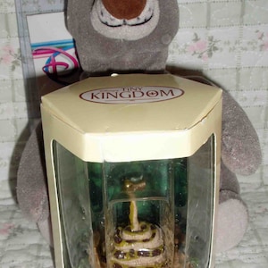 May include: A plush gray bear toy with a brown nose and a smiling face. In front of the bear is a clear display case containing a miniature statue of a snake. The display case has the words "Tiny Kingdom" printed on it.