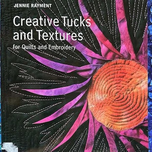 May include: Book cover with the title "Creative Tucks and Textures for Quilts and Embroidery" in black text on a black background. The cover features a close-up of a colorful fabric flower with pink, purple, and orange hues.