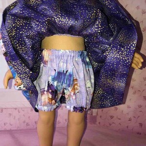 K&ouml;nnte beinhalten: Puppenkleidung-Set mit einem lila Umhang mit goldenen Sprenkeln und passenden Shorts. Die Shorts haben einen hellvioletten Hintergrund mit einem Cartoon-Print. Das Outfit der Puppe ist f&uuml;r ein Kinderspielzeug bestimmt.