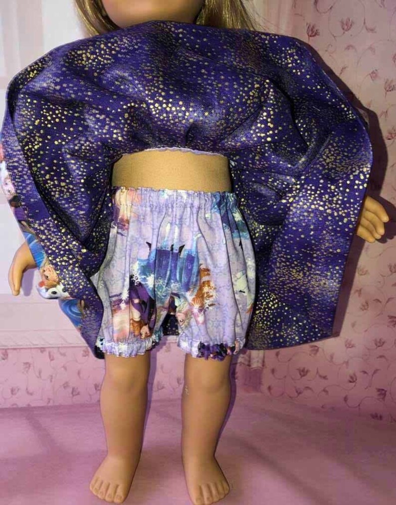 K&ouml;nnte beinhalten: Puppenkleidung-Set mit einem lila Umhang mit goldenen Sprenkeln und passenden Shorts. Die Shorts haben einen hellvioletten Hintergrund mit einem Cartoon-Print. Das Outfit der Puppe ist f&uuml;r ein Kinderspielzeug bestimmt.