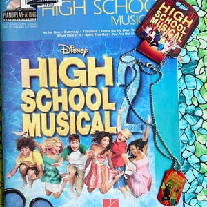 May include: A High School Musical 2 piano play-along book with a matching dog tag necklace. The book cover is blue with the movie title in large gold letters and features images of the cast. The dog tag is red with a silver chain.
