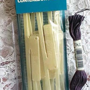 May include: A package of 10 DMC Stitch Bow floss holders, also known as thread organizers or archs. The package is blue and white with the DMC logo and the text "Stitch Bow Floss Holder Archets Contenedor de Hilo". The package is next to a skein of purple embroidery floss.
