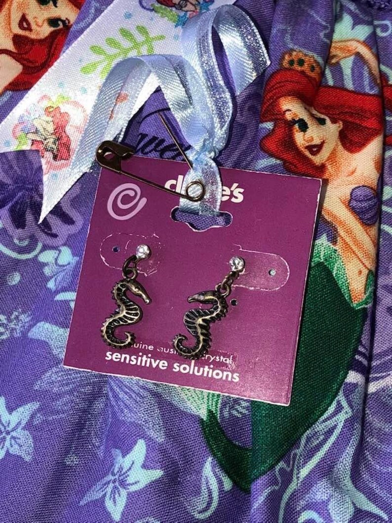 Op de afbeelding: Een paar zeepaardjesoorbellen op een paarse kaart met het Claire's logo. De oorbellen zijn van donker metaal met een kristallen accent. De kaart is bevestigd aan een blauw lint met een veiligheidsspeld. De achtergrond is een paarse stof met een zeemeerminprint.