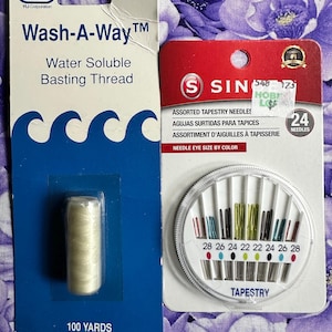 May include: Two sewing supplies are displayed against a purple floral background. On the left is a package of "Wash-A-Way" water-soluble basting thread, 100 yards. On the right is a package of 24 assorted tapestry needles, with needle eye size indicated by color.