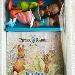 May include: A boxed set featuring Peter Rabbit figurines and a book titled "The World of Peter Rabbit" by Beatrix Potter. The book cover displays an illustration of Peter Rabbit and a chipmunk in a garden setting. The figurines are in various colors.