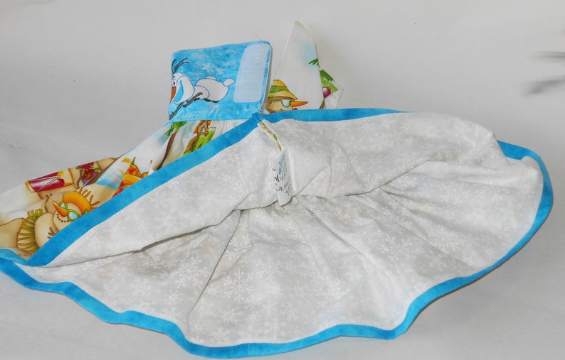 Pode incluir: Um vestido branco com uma bainha azul e uma estampa colorida com personagens do filme Frozen. A estampa inclui Olaf, Elsa e Anna.