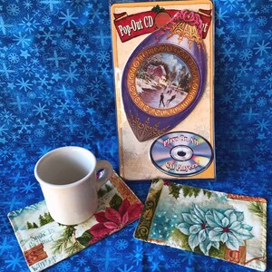 May include: Two Christmas-themed fabric coasters with a winter scene and a poinsettia design. The coasters are on a blue background with white snowflakes. A white ceramic mug is sitting on one of the coasters. A CD case with a pop-out CD is in the background. The CD case has a winter scene with a house and people ice skating. The text on the CD case reads "Pop-Out CD" and "Plays On All CD Players."