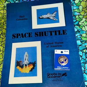 May include: A blue book titled "SPACE SHUTTLE" with images of the space shuttle. A pin with a space shuttle design is on the book. The book has the text "United States of America" and "Graphs by Goodman".