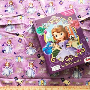 May include: A purple book with a picture of Princess Sofia the First on the cover. The book is titled "Sofia the First" and "My Busy Books". The book is lying on a purple fabric with a repeating pattern of Princess Sofia the First.