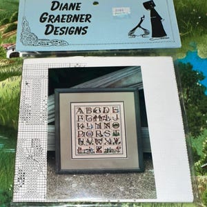 May include: A cross-stitch pattern by Diane Graebner Designs. The pattern features an alphabet with decorative images. The pattern is framed and packaged with a light blue header that includes the designer's name and logo.