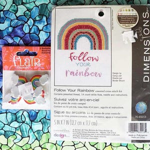 May include: A craft kit featuring a rainbow cross-stitch design with the text "Follow Your Rainbow." The kit includes pre-sorted thread, a 14-count white Aida cloth, a needle, and instructions. Also included are Flair Originals rainbow and cloud buttons.