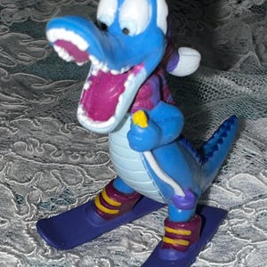 May include: A blue cartoon dinosaur figurine on skis. The dinosaur has a white head, a purple mouth, and is wearing a purple and red scarf. The skis are purple with yellow and red accents. The figurine is posed as if skiing.
