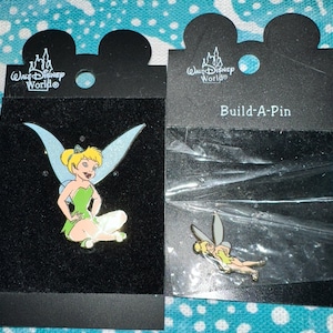 May include: Two Disney World Build-A-Pin pins featuring Tinkerbell. One pin is displayed on a black backing, showing Tinkerbell in a green dress and light blue wings. The other pin is in a plastic bag.