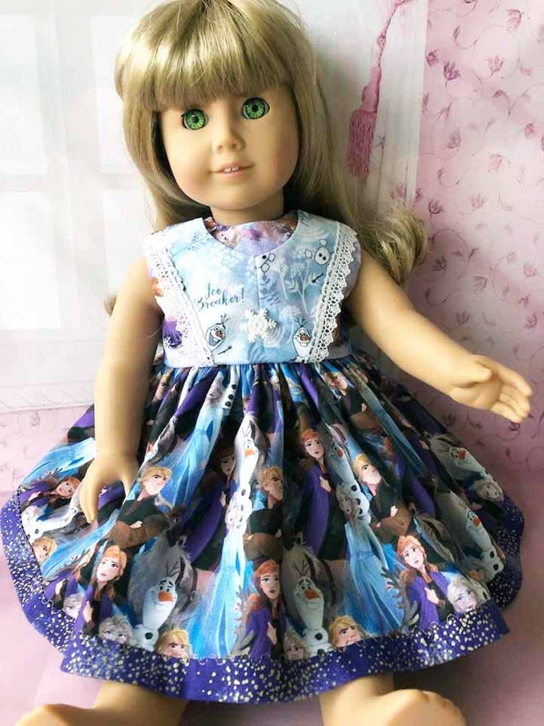 K&ouml;nnte beinhalten: Eine Puppe in einem blau-violetten Kleid mit Frozen-Motiv. Das Kleid zeigt Charaktere aus dem Film, darunter Elsa und Olaf. Das Oberteil hat den Text "Ice Breaker!" und eine wei&szlig;e Spitzenborte.