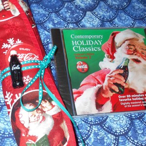 May include: A red and white Christmas stocking with a green ribbon and a miniature Coca-Cola bottle. The stocking is rolled up and tied with a green ribbon. A CD case with a green background and a picture of Santa Claus holding a Coca-Cola bottle is in the background. The CD case has the text "Contemporary Holiday Classics Volume 2" on it.
