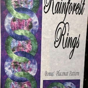 May include: A fabric panel with the text "Rainforest Rings" in elegant script. The design features interlocking circles in shades of green, blue, and purple, with a floral pattern in the center. Includes a bonus placemat pattern.