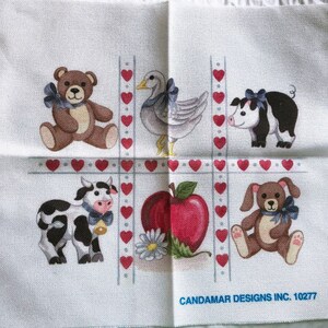 May include: White fabric square featuring a tic-tac-toe grid with farm animal illustrations. A brown teddy bear, a gray goose, a black and white pig, a black and white cow, a red apple, and a brown bunny are depicted.