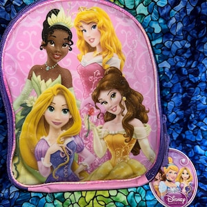 May include: A pink backpack featuring four Disney Princesses: Tiana, Aurora, Belle, and Rapunzel. The backpack has a purple zipper and trim, with a small Disney Princesses tag attached. The background is a colorful, patterned fabric.