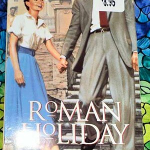 May include: A book cover for the movie "Roman Holiday" featuring Gregory Peck and Audrey Hepburn. The cover shows the actors holding hands, with Hepburn in a white blouse and blue skirt. The title is in large, white letters.