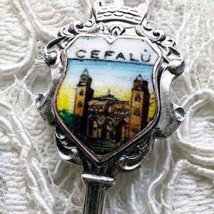 May include: Silver souvenir spoon with a colorful enamel design of a building with a crown on top. The spoon has the word "CEFALU" written on it.
