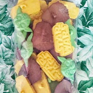 May include: A clear plastic bag filled with Star Wars-themed buttons. The buttons are yellow, green, and brown, featuring characters and logos from the franchise. The bag has a header with the Star Wars logo and Darth Vader.
