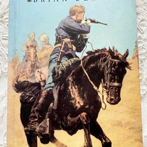 May include: Book cover for "Soldier Boy" by Brian Burks. The cover art depicts a soldier on horseback aiming a rifle, with other riders in the background. The title is in a bold, stylized font against a light blue sky.
