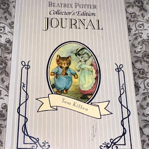 May include: A Beatrix Potter Collector's Edition journal with a light gray cover and vertical stripes. The cover features an oval illustration of two anthropomorphic cats and the text "JOURNAL" in large letters. The journal is titled "Tom Kitten".