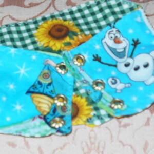 May include: A blue, green, and white fabric scrap with a patchwork design. The fabric features a green gingham pattern, a sunflower, and a cartoon image of Olaf from the movie Frozen. The fabric also has a blue background with white stars and a cartoon image of a princess.