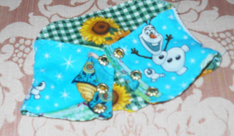 May include: A blue, green, and white fabric scrap with a patchwork design. The fabric features a green gingham pattern, a sunflower, and a cartoon image of Olaf from the movie Frozen. The fabric also has a blue background with white stars and a cartoon image of a princess.