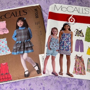 May include: Two vintage McCall's sewing pattern envelopes. One features a girl in a plaid dress, the other shows a girl in a floral dress. The patterns are for girls' clothing, including dresses and tops.