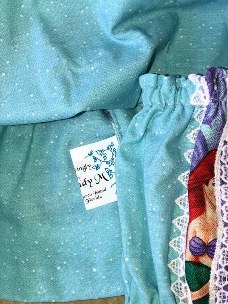 Op de afbeelding: Close-up van lichtblauwe stof met witte stippen. De stof is gerimpeld en onthult een label met de tekst "ingly M" en "Marco Island Florida". Een gedeelte van de stof met een zeemeerminillustratie en witte kanten rand is zichtbaar.