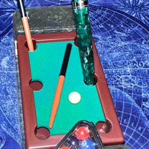 May include: A miniature pool table set with a green felt surface, brown frame, and a white cue ball. Includes a triangle rack of colorful pool balls, two cues, and a green and black marbled pen.