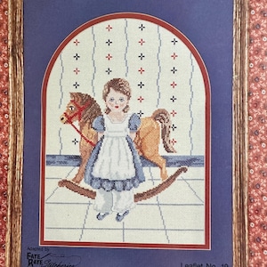 May include: Framed cross-stitch artwork featuring a young girl in a blue dress and white apron, standing next to a rocking horse. The design is set against a blue background with a patterned archway. The frame is brown, and the artwork is labeled "Leaflet No. 19".