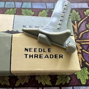 May include: A white box with the words "NEEDLE THREADER" printed on it. A gray plastic needle threader with the word "KENMORE" printed on it is on top of the box.